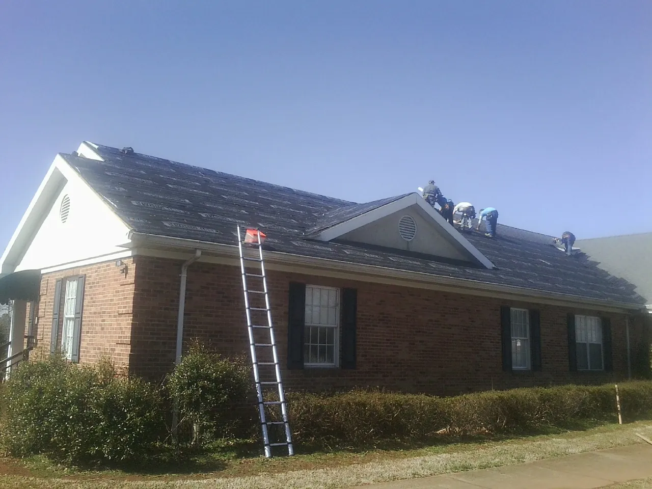 Crew tearing shingles off a brick ranch roof with a ladder set — Emergency Roof Repair work by our crew in Pine Ridge